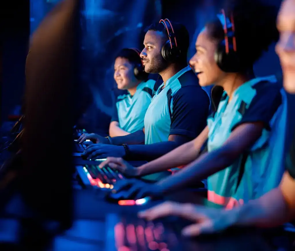 Zespół graczy e-sportowych siedzi obok siebie przy komputerach do gier podczas meczu. Noszą słuchawki i skupiają się na grze w oświetlonej arenie e-sportowej.